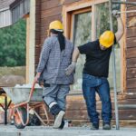 Workers comp benefits (Two construction workers wearing hard hats work at an outdoor site, one with a wheelbarrow.)