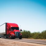 18 wheeler accident lawyer (Vibrant red semi-trailer truck driving on an open highway under a clear blue sky.)