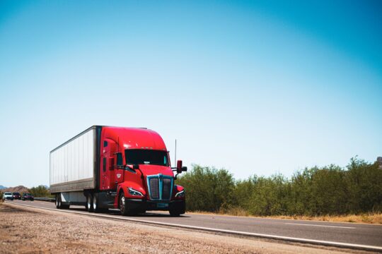 18 wheeler accident lawyer (Vibrant red semi-trailer truck driving on an open highway under a clear blue sky.)